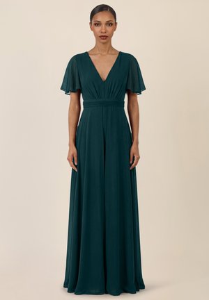 Frau in einem bodenlangen dunkelgrünen Kleid mit flatternden Ärmeln und tiefem V-Ausschnitt, stehend vor einem schlichten beigefarbenen Hintergrund.
