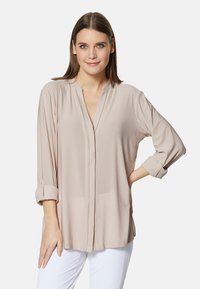 Helle beige Bluse mit langen Ärmeln, V-Ausschnitt, hochgerollten Bündchen und einem feinen Nahtdetail vorne. Hergestellt aus weichem, glattem Stoff.