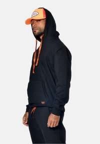 Sweat à capuche noir avec des cordons orange et un logo, associé à un jogging noir assorti. La casquette orange arbore un badge logo sur le devant.
