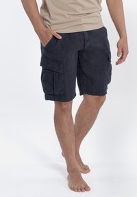 Pantaloni cargo neri con più tasche laterali, in cotone, con una vestibilità comoda e orlo poco sopra il ginocchio. Indossati a piedi nudi.