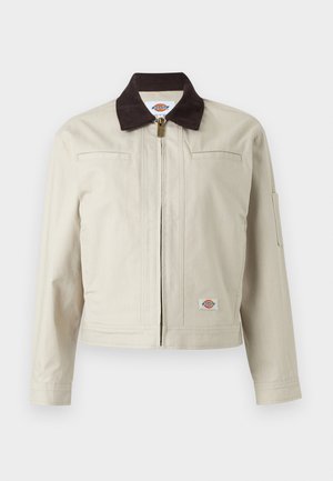 Beige jakke med glidelås, mørk brun krage, brystlommer og logoemblem på nedre kant, vist mot ensfarget bakgrunn.