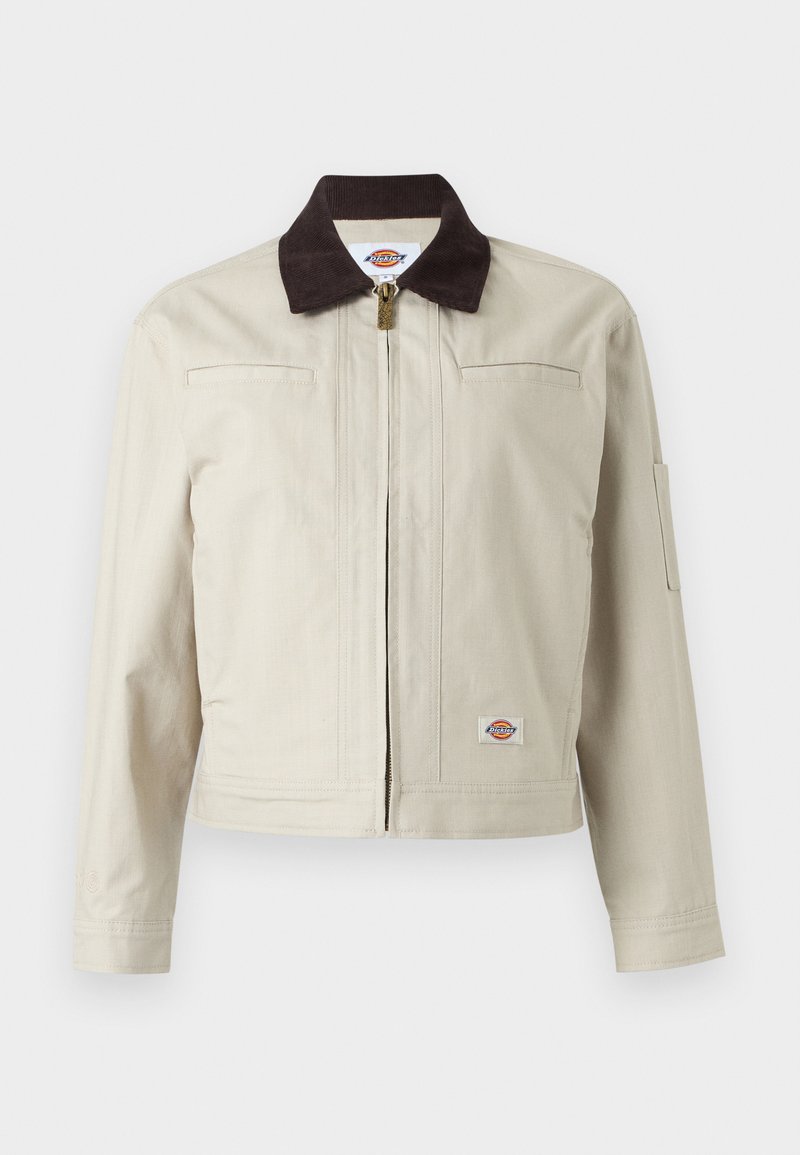 Beige Reißverschlussjacke mit dunkelbraunem Kragen, Brusttaschen und Logoaufnäher am unteren Saum, vor einfarbigem Hintergrund präsentiert.