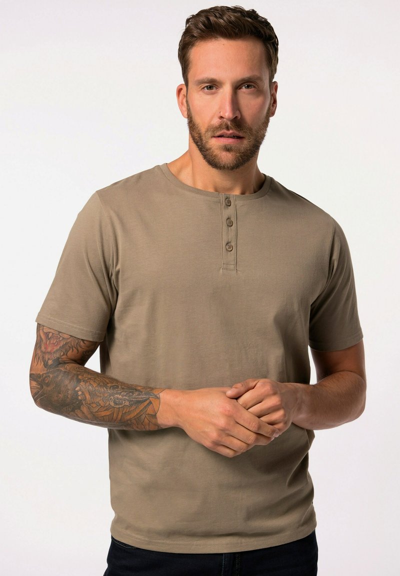 Beżowa koszulka henley z krótkim rękawem, z trzema guzikami przy dekolcie, wykonana z miękkiego materiału; model z tatuażami na jednym ramieniu.