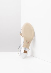 Sandale en cuir argenté avec un talon bas, présentant un bout arrondi et une semelle en caoutchouc beige. Comprend un marquage de la marque et une étiquette de taille.