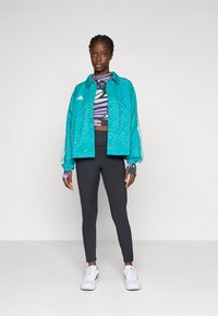 Fată tânără în picioare, purtând o jachetă cu model teal, un top scurt cu model colorat, colanți negri și adidași albi, pe un fundal simplu.