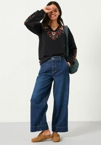 Schwarzer Sweatshirt mit buntem floralem Stickerei am Rundhalsausschnitt und den Ärmeln, kombiniert mit weit geschnittenen blauen Jeans und braunen Holzclogs.