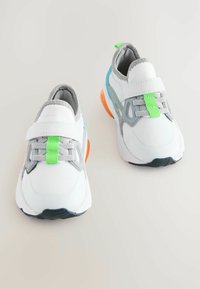 Weiße Sportschuhe mit grauen Akzenten, aus Mesh-Material, orangefarbenem Absatz und grünen Details an Riemen und Schnürsenkeln. Abgerundete Zehenform.