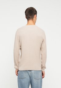Suéter de punto color beige claro con un patrón texturizado, puños y dobladillo acanalados. Cuenta con un cuello redondo y un corte relajado, usado sobre jeans azules.