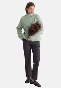Grüner Pullover mit Zopfmuster, breite Bündchen, kombiniert mit dunkelgrauer Hose. Hält eine braune Pelz-Clutch, schwarze Schuhe mit strukturiertem Design.
