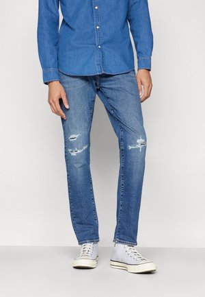 Lyseblå denimjeans med rett snitt og slitte detaljer. Kombinert med en blå skjorte med knapper og lysegrå sneakers.