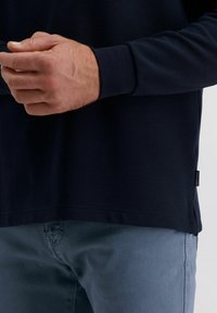 Mann trägt ein marineblaues, geripptes Langarmshirt und eine blaue Hose, die Hände vor der Brust gefaltet, steht vor einem neutralen Hintergrund.