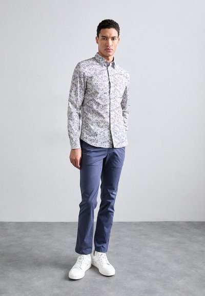 Camicia button-up con piccolo motivo floreale in bianco, blu e viola, abbinata a pantaloni navy e sneakers bianche, in piedi su un pavimento grigio.