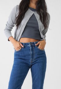 Cardigan grigio a coste con bottoni sopra un top corto grigio scuro, abbinato a jeans in denim blu a vita alta con tasche frontali.
