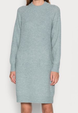 Femme portant une robe-pull tricotée bleu clair, manches longues et longueur genou, avec un col rond, sur un fond uni.