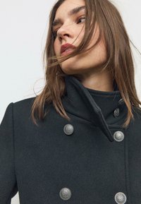 Cappotto nero doppiopetto realizzato in tessuto strutturato, presenta un colletto alto e bottoni decorativi argentati con design intricati.