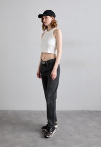 Zwarte cap, witte cropped tanktop en donkere jeans. Zwarte sneakers met zilveren accenten. Model poseert zijwaarts tegen een neutrale achtergrond.