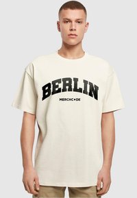Kremkrāsas pārspīlēts t-krekls ar drosmīgu melnu tekstu "BERLĪNE" un "MERCH•DE" arkas dizainā. Īsas piedurknes, maiga tekstūra.