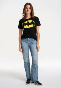 Black cotton t-shirt with a large yellow Batman logo on the front, paired with light blue straight-leg jeans and black sneakers.