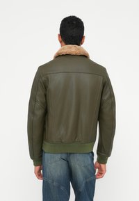Uomo che indossa un bomber verde oliva con collo in shearling beige e jeans blu, in piedi con la schiena rivolta verso uno sfondo bianco semplice.