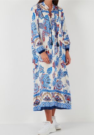 Longue robe blanche avec des motifs floraux bleus et rouges, manches longues avec des poignets bleus, et des baskets blanches sur une personne debout.