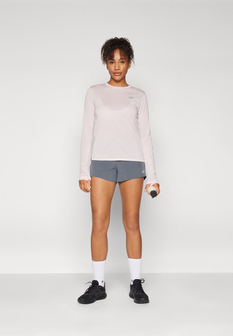 Hellrosa langärmliges Sportshirt, graue Shorts, schwarze Turnschuhe, weiße Knöchelsocken, hält eine Hantel, vor einem schlichten weißen Hintergrund.