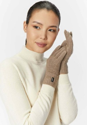 Frau in cremefarbenem Rollkragenpullover, die braune gestrickte Handschuhe mit einem kleinen schwarzen Etikett trägt und anpasst, vor weißem Hintergrund.