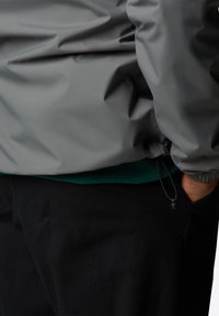 Grijze lichtgewicht jas met een elastische zoom, gelaagd over een groene geribbelde sweatshirt, gecombineerd met zwarte broek. Zachte textuur en casual ontwerp.