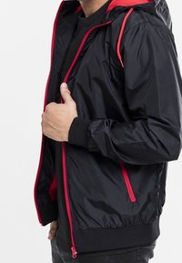 Schwarze Kapuzenjacke mit roten Akzenten, Reißverschluss vorne und Rippbündchen. Hergestellt aus einem glänzenden Stoff mit glatter Textur.