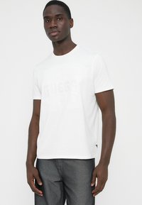 T-shirt bianca in cotone con un delicato logo in rilievo sul davanti, scollo rotondo e maniche corte, abbinata a jeans in denim grigio.