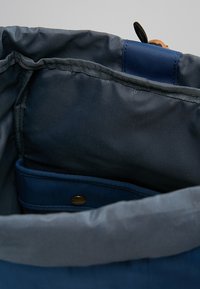 Interno di una borsa di stoffa blu con una trama liscia. Presenta una tasca e un dettaglio con pulsante dorato. I bordi sono cuciti, con una fodera blu scuro.