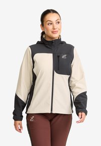 Woman wearing a beige and black zip-up jacket with a high collar and brown pants, looking to the side with a neutral expression.