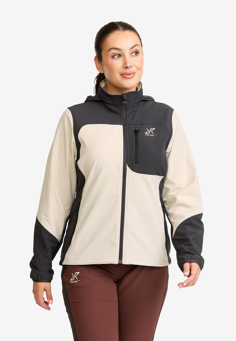 Woman wearing a beige and black zip-up jacket with a high collar and brown pants, looking to the side with a neutral expression.