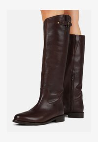 Botas altas de cuero marrón hasta la rodilla con punta redonda, cremallera lateral y un pequeño detalle de hebilla en la parte superior, con una textura suave.