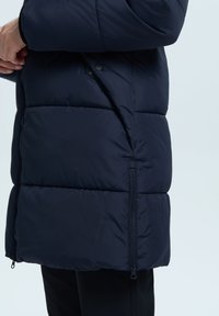 Marineblauwe pufferjack met gestikte secties, zichtbare zijrits en een soepele textuur, voorzien van een staande kraag en drukknoopsluiting.
