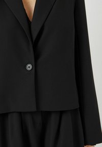 Černý blazer s jedním knoflíkem, strukturovaný design a hladký povrch. Má elegantní klopu a zkrácenou délku.