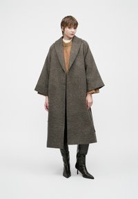 Woman wearing a long brown patterned coat over an orange sweater and black high-heeled boots, standing against a white background.