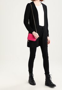 Black long cardigan over a white ribbed top, paired with black skinny pants and lace-up black boots. Accessories include a small pink crossbody bag.