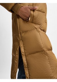 Bruine pufferjas met een gewatteerd ontwerp, voorzien van een zijrits en ribgebreide manchetten. De stof is glad en glanzend, met een zichtbare zak.