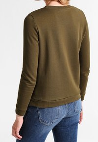 Persona vestida con una camisa de manga larga verde oliva y jeans de denim azules, vista desde la espalda con la mano izquierda relajada a un lado.