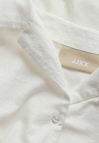 Λευκό πουκάμισο με υφή και γιακά με εμφανές μπεζ ετικέτα της μάρκας JJXX μέσα στον λαιμό και ένα μόνο λευκό κουμπί κουμπωμένο.