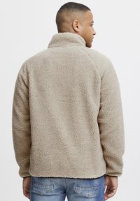 Beige fleece jacket with a high collar, textured fabric, and long sleeves. Features a relaxed fit and no visible closures.