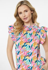 Bluse mit floralem Muster und Rüschensäumen, in Farben wie Pink, Orange und Grün auf einem hellen Hintergrund. Rüschendetail am Halsausschnitt.