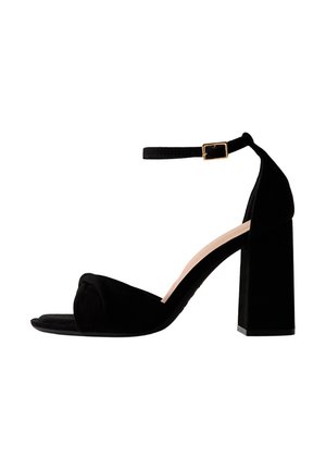 Zwarte suède sandaal met hoge hak en blokhak, open teen en enkelbandje met gouden gesp, op een witte achtergrond.