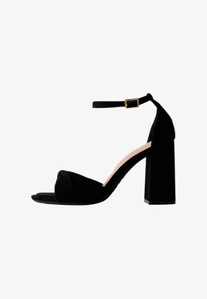 Sandalo con tacco alto in camoscio nero, con tacco largo, punta aperta e cinturino alla caviglia con fibbia dorata su sfondo bianco.