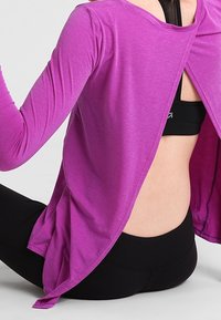 Lila långärmad topp med en öppen ryggdesign, tillverkad av ett lätt material, kombinerad med en svart sport-bh och leggings.