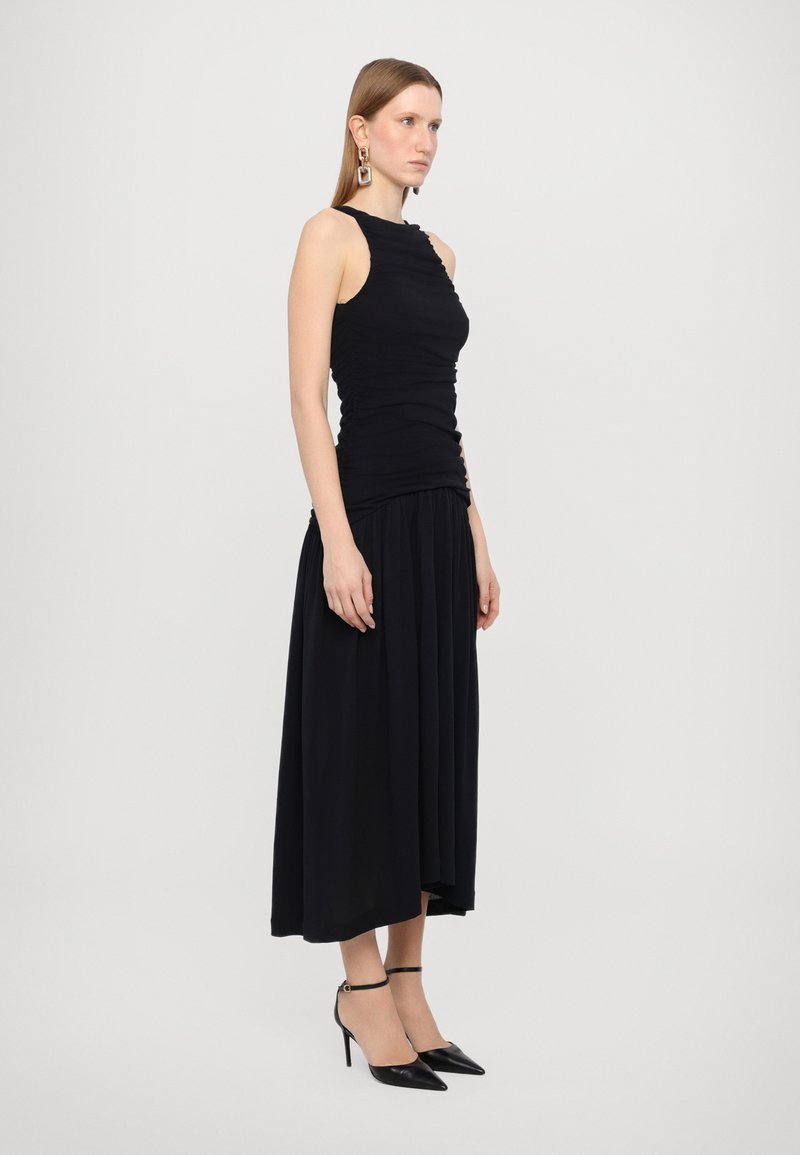 Schwarzes Kleid mit tailliertem, gerafftem Oberteil und fließendem Rock. Das Model kombinierte es mit schwarzen spitzen Absatzschuhen und kleinen Creolen.