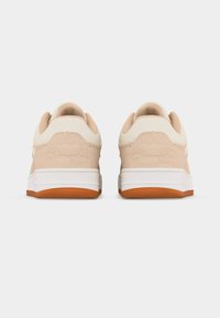 Ζευγάρι μπεζ και λευκά αθλητικά παπούτσια με πορτοκαλί λαστιχένιες σόλες, εμφανιζόμενα από πίσω σε απλό γκρι φόντο.