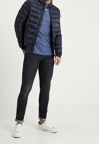 Granatowa kurtka puchowa z pikowanym wykończeniem, niebieska koszulka polo, czarne slim fit dżinsy oraz białe sneakersy, z podwiniętymi rękawami i zamkiem błyskawicznym.