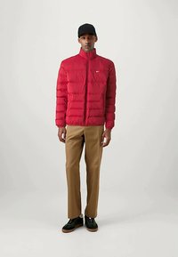 Mann kledd i en rød dunjakke, brune bukser, svarte joggesko og svart caps, som står mot en ensfarget lys bakgrunn.