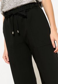 Črne elastične hlače z vrvico, ki imajo srebrne kovinske detajle in stranske žepke, narejene iz gladkega, lahkega materiala.